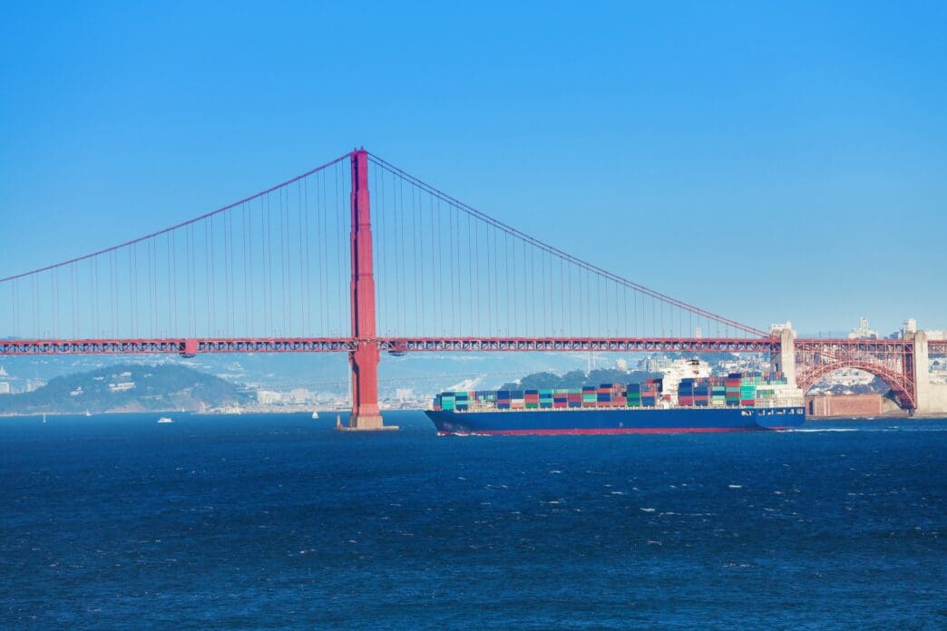 The iconic Golden Gate Bridge, a large red suspension bridge, spans across a blue body of water. A large container ship, laden with colorful cargo, sails directly underneath the bridge's impressive structure.