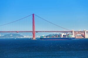 The iconic Golden Gate Bridge, a large red suspension bridge, spans across a blue body of water. A large container ship, laden with colorful cargo, sails directly underneath the bridge's impressive structure.