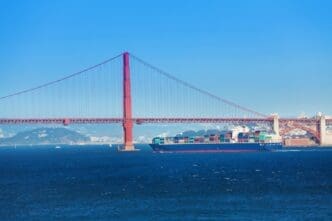 The iconic Golden Gate Bridge, a large red suspension bridge, spans across a blue body of water. A large container ship, laden with colorful cargo, sails directly underneath the bridge's impressive structure.