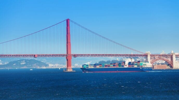The iconic Golden Gate Bridge, a large red suspension bridge, spans across a blue body of water. A large container ship, laden with colorful cargo, sails directly underneath the bridge's impressive structure.