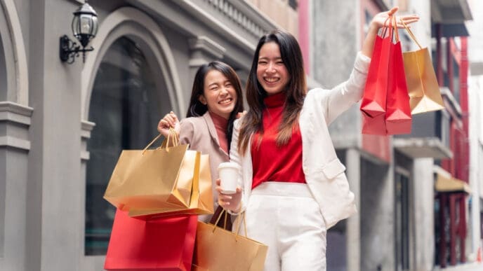 Two happy young women in China, are smiling and laughing while walking down a Chinese city shopping street, holding multiple red and gold shopping bags.