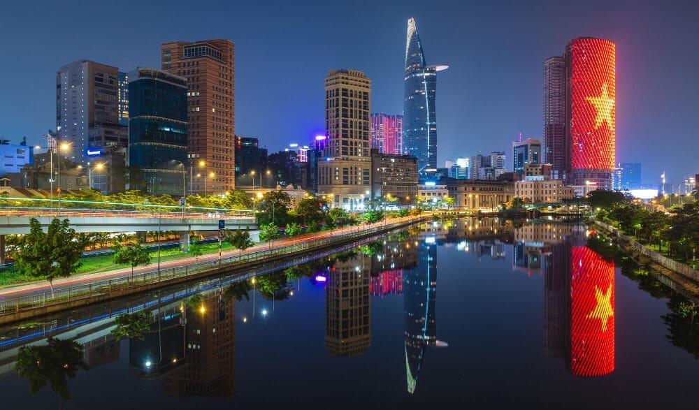 A vibrant nighttime cityscape of Ho Chi Minh City, Vietnam, showing modern skyscrapers and illuminated buildings, including one lit up with the Vietnamese flag, all perfectly reflected in a calm river.