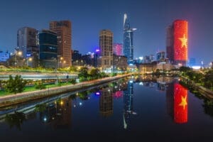 A vibrant nighttime cityscape of Ho Chi Minh City, Vietnam, showing modern skyscrapers and illuminated buildings, including one lit up with the Vietnamese flag, all perfectly reflected in a calm river.
