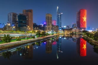 A vibrant nighttime cityscape of Ho Chi Minh City, Vietnam, showing modern skyscrapers and illuminated buildings, including one lit up with the Vietnamese flag, all perfectly reflected in a calm river.