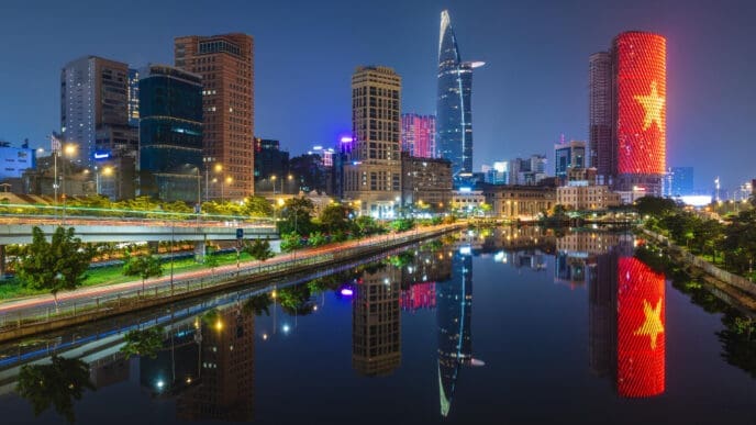A vibrant nighttime cityscape of Ho Chi Minh City, Vietnam, showing modern skyscrapers and illuminated buildings, including one lit up with the Vietnamese flag, all perfectly reflected in a calm river.
