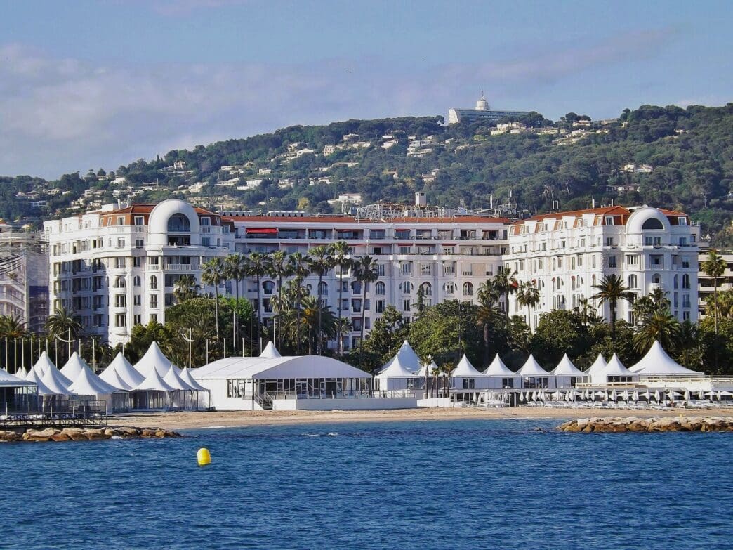 The grand, white facade of the Hotel Barrière Le Majestic Cannes, with its reddish-brown roof, stands on a sandy beach. Numerous white event tents are set up on the sand, overlooking the blue sea.