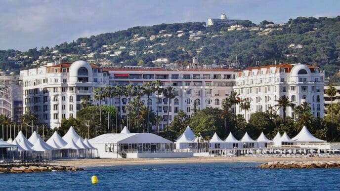 The grand, white facade of the Hotel Barrière Le Majestic Cannes, with its reddish-brown roof, stands on a sandy beach. Numerous white event tents are set up on the sand, overlooking the blue sea.