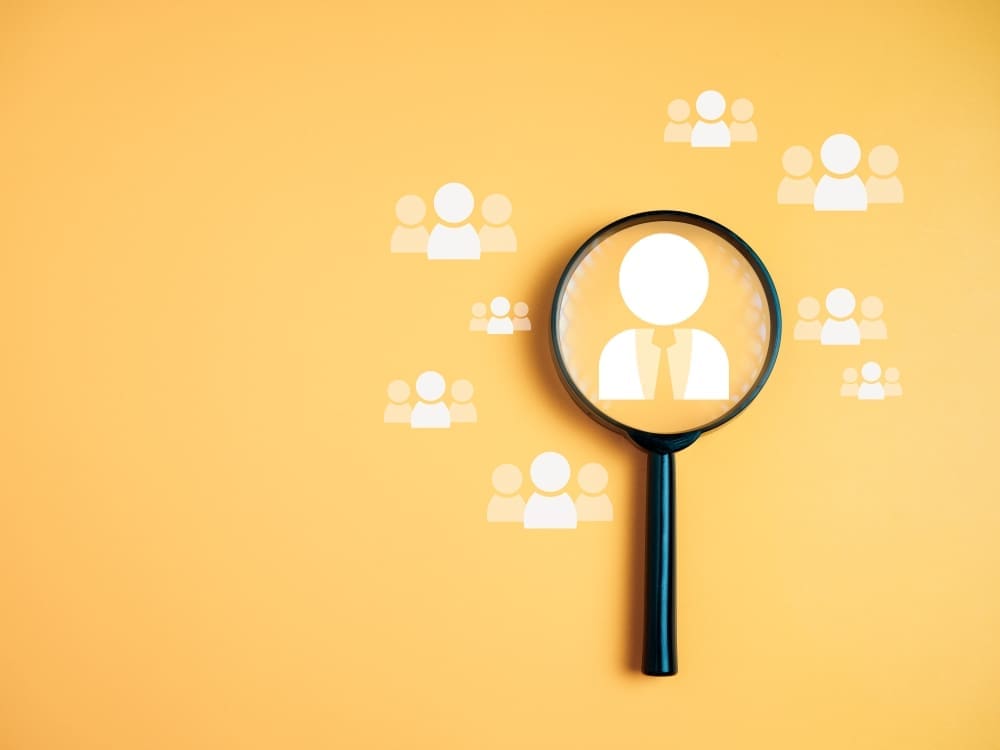 A magnifying glass on a yellow background is positioned over a white icon of a person in a tie, with other people icons scattered around it, symbolizing the process of recruitment and human resources.