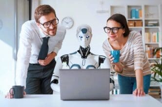 A humanoid robot sits at a desk with a laptop, flanked by a male and female colleague who are both leaning in to look at the screen, symbolizing human-robot collaboration in a modern office.