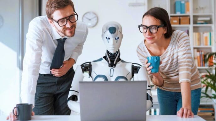 A humanoid robot sits at a desk with a laptop, flanked by a male and female colleague who are both leaning in to look at the screen, symbolizing human-robot collaboration in a modern office.