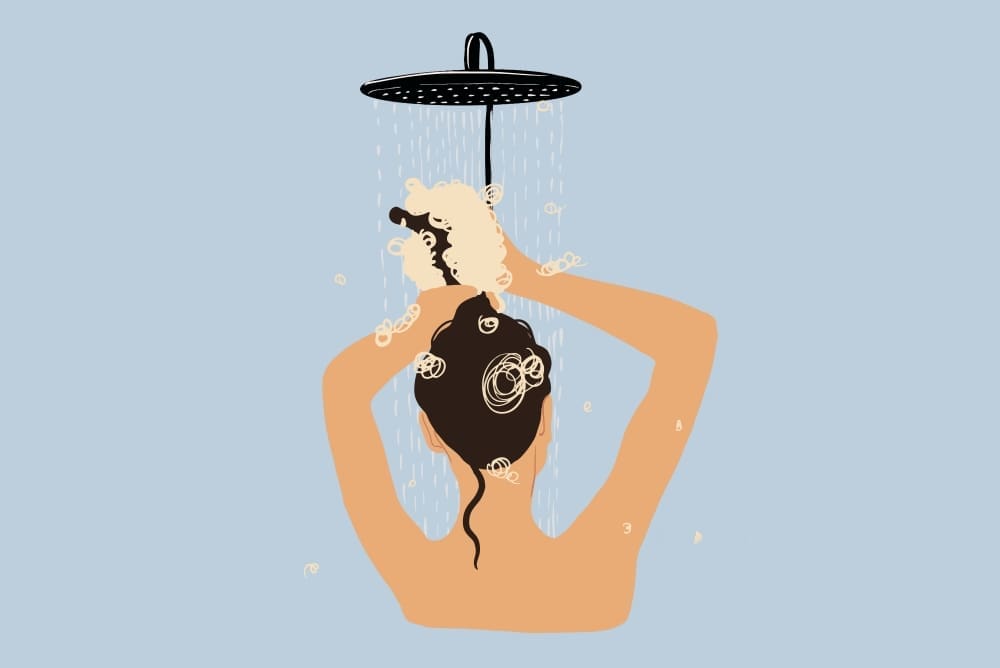 A stylized illustration of a person with dark hair seen from behind, washing their hair with shampoo lather under a shower head against a light blue background.