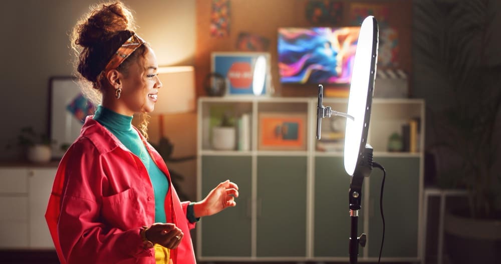 A smiling young woman, who appears to be Black or of mixed-race descent, stands in front of a ring light and smartphone, confidently speaking as she records content for an audience.