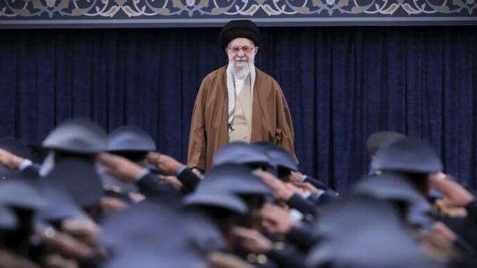 Iran's Supreme Leader Ayatollah Ali Khamenei stands in front of a blue curtain, looking at a group of uniformed individuals in the foreground who are saluting him.