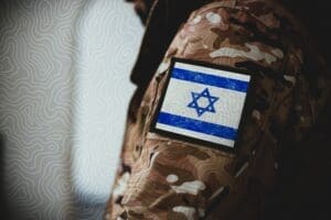 A close-up photograph of a soldier's uniform sleeve, which has a desert camouflage pattern and features a patch of the national flag of Israel. A white topographic map pattern is overlaid on the left side of the image.