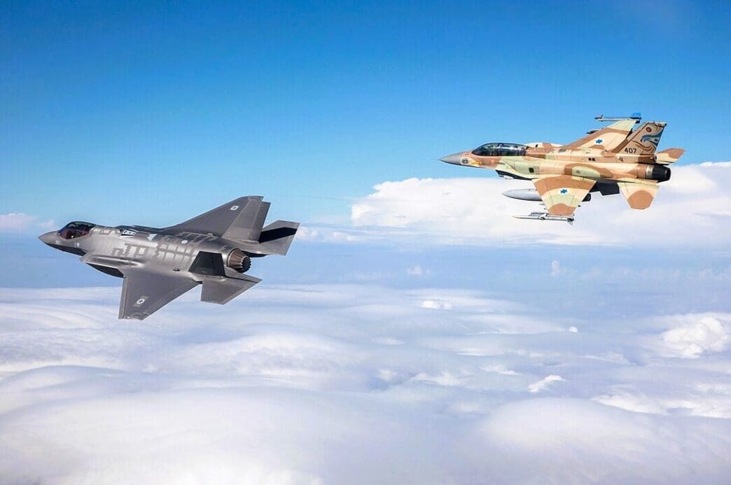 An Israeli Air Force F-35I Adir stealth fighter jet and an F-16I Sufa fighter jet in desert camouflage, flying together in formation high above a layer of white clouds against a blue sky.