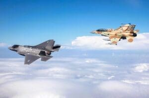 An Israeli Air Force F-35I Adir stealth fighter jet and an F-16I Sufa fighter jet in desert camouflage, flying together in formation high above a layer of white clouds against a blue sky.