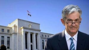 A composite image showing Federal Reserve Chairman Jerome Powell on the right, superimposed next to a photograph of the neoclassical Federal Reserve Board Building in Washington, D.C., on the left, under a clear blue sky.