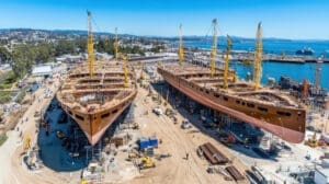 An aerial view of a bustling shipyard showing two massive ship hulls, appearing to be for large sailing vessels, under construction. They are surrounded by scaffolding, numerous yellow cranes, and workers, with a busy port visible in the background.