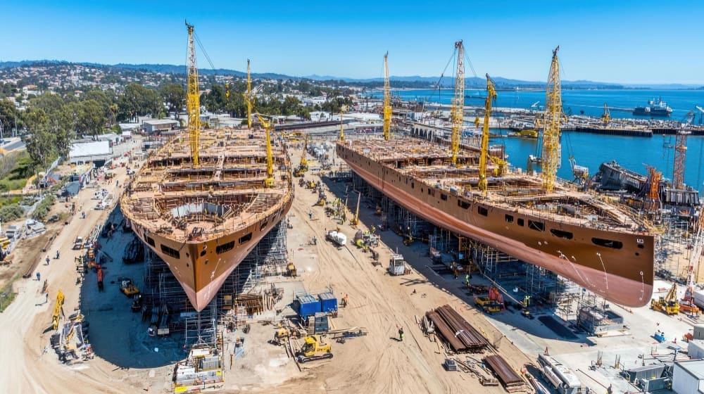 An aerial view of a bustling shipyard showing two massive ship hulls, appearing to be for large sailing vessels, under construction. They are surrounded by scaffolding, numerous yellow cranes, and workers, with a busy port visible in the background.