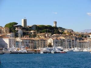 A sunny daytime view of the historic old town of Cannes, France, with traditional buildings climbing a hillside. Two prominent towers overlook a harbor filled with numerous yachts and sailboats.