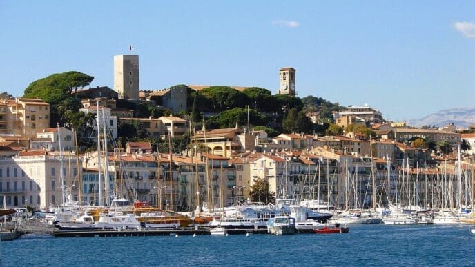 A sunny daytime view of the historic old town of Cannes, France, with traditional buildings climbing a hillside. Two prominent towers overlook a harbor filled with numerous yachts and sailboats.