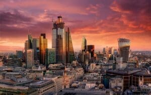 The modern skyscrapers of the London financial district, such as the Gherkin and the Walkie-Talkie building, are silhouetted against a dramatic and colorful sunset sky.