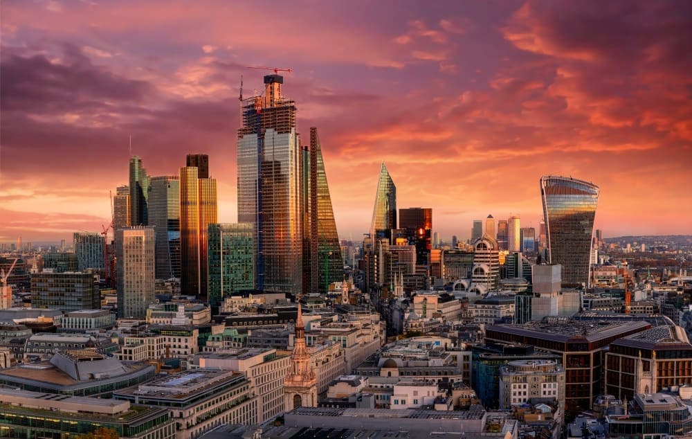 The modern skyscrapers of the London financial district, such as the Gherkin and the Walkie-Talkie building, are silhouetted against a dramatic and colorful sunset sky.
