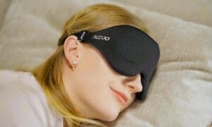 A woman with blonde hair is shown sleeping peacefully on her side while wearing a black, contoured MZOO 3D sleep eye mask that covers her eyes.