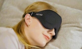 A woman with blonde hair is shown sleeping peacefully on her side while wearing a black, contoured MZOO 3D sleep eye mask that covers her eyes.