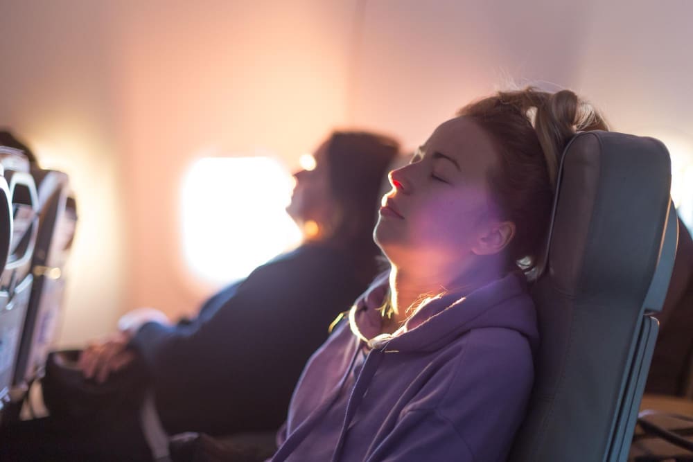 A young woman who appears to be White, with her eyes closed and head tilted back, sits peacefully in an airplane seat, illuminated by warm sunlight from a window, suggesting meditation or deep relaxation.