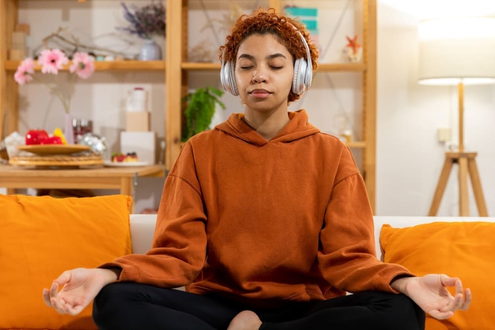 A young woman who appears to be Black or mixed-race, with short curly hair, sits cross-legged on a sofa with her eyes closed, wearing white over-ear headphones and holding her hands in a meditation mudra.