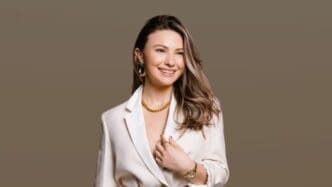 A professional studio portrait of entrepreneur Mimi Bouchard, smiling while looking off-camera. She has long brown hair and is wearing a white blazer with gold jewelry against a solid taupe background.