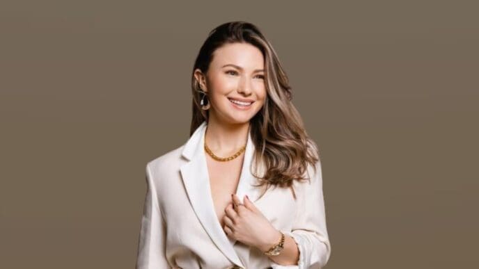 A professional studio portrait of entrepreneur Mimi Bouchard, smiling while looking off-camera. She has long brown hair and is wearing a white blazer with gold jewelry against a solid taupe background.