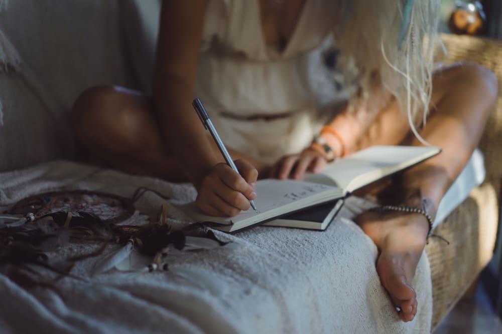 A happy young woman sits in a cozy, comfortable room, smiling as she focuses on writing or drawing in a notebook.