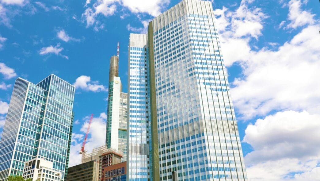 A low-angle view looking up at several modern skyscrapers with glass and metal facades, set against a blue sky with white clouds, typical of a major financial district.