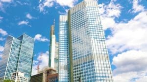 A low-angle view looking up at several modern skyscrapers with glass and metal facades, set against a blue sky with white clouds, typical of a major financial district.