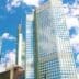 A low-angle view looking up at several modern skyscrapers with glass and metal facades, set against a blue sky with white clouds, typical of a major financial district.