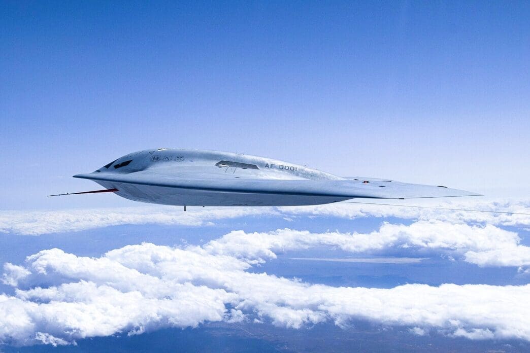 A light grey Northrop Grumman B-21 Raider stealth bomber flies high in the sky above a layer of white clouds, showcasing its sleek, next-generation "flying wing" design.