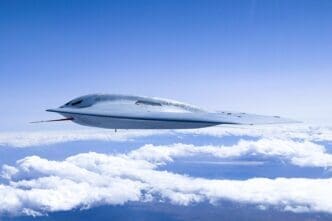 A light grey Northrop Grumman B-21 Raider stealth bomber flies high in the sky above a layer of white clouds, showcasing its sleek, next-generation "flying wing" design.