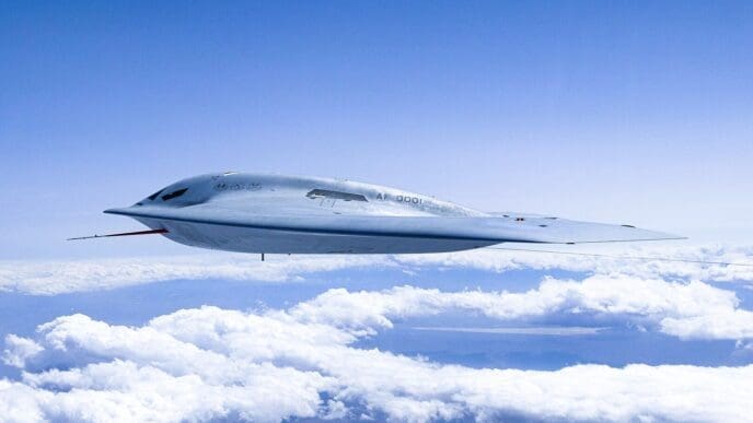 A light grey Northrop Grumman B-21 Raider stealth bomber flies high in the sky above a layer of white clouds, showcasing its sleek, next-generation "flying wing" design.
