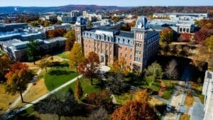 An aerial view of the historic Old Main building at the University of Arkansas. The large brick structure, with its prominent towers and clock faces, is surrounded by green lawns and trees with vibrant autumn foliage under a clear blue sky.