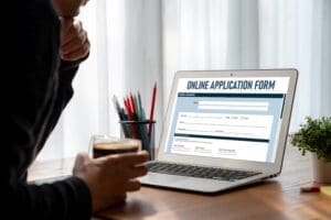 A person views a laptop screen displaying a blank "ONLINE APPLICATION FORM," with a glass of coffee and pens on the desk in an office or home office setting.
