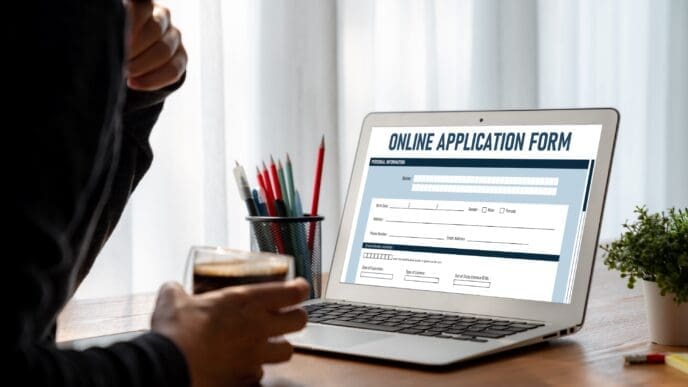A person views a laptop screen displaying a blank "ONLINE APPLICATION FORM," with a glass of coffee and pens on the desk in an office or home office setting.
