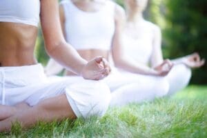 Three women in white athletic wear sit in a cross-legged meditation pose on green grass, with their hands in a mudra. The foreground is in sharp focus, while other meditators are blurred in the background.