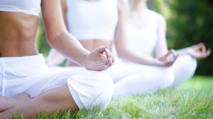 Three women in white athletic wear sit in a cross-legged meditation pose on green grass, with their hands in a mudra. The foreground is in sharp focus, while other meditators are blurred in the background.