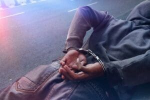 A person wearing a hooded sweatshirt is lying on the pavement with their hands restrained behind their back in handcuffs, with what appear to be police lights glowing on them.