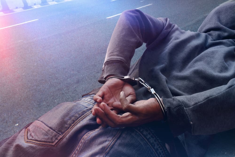 A person wearing a hooded sweatshirt is lying on the pavement with their hands restrained behind their back in handcuffs, with what appear to be police lights glowing on them.
