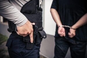 A police officer rests a hand on their holstered handgun while standing next to a suspect who has their hands behind their back in handcuffs.