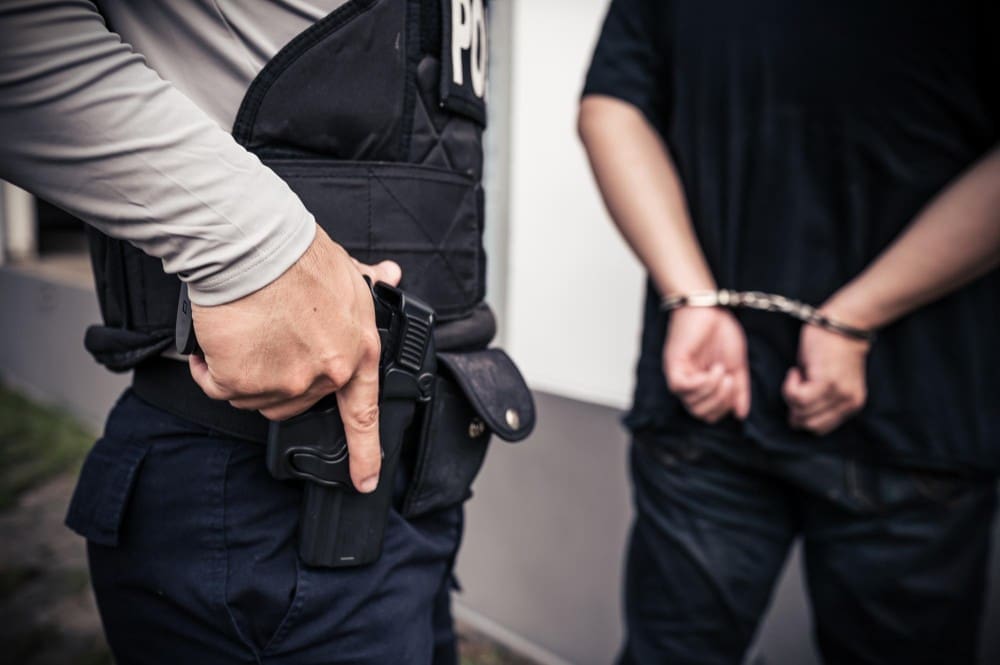 A police officer rests a hand on their holstered handgun while standing next to a suspect who has their hands behind their back in handcuffs.