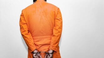 A person wearing a bright orange jumpsuit is seen from behind, with their hands cuffed behind their back, standing against a plain light-colored background.